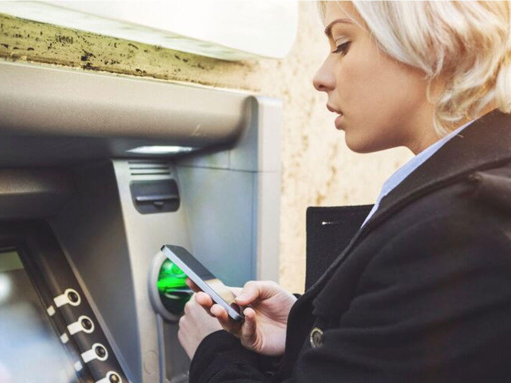 Woman using her smartphone in front of an ATM machine